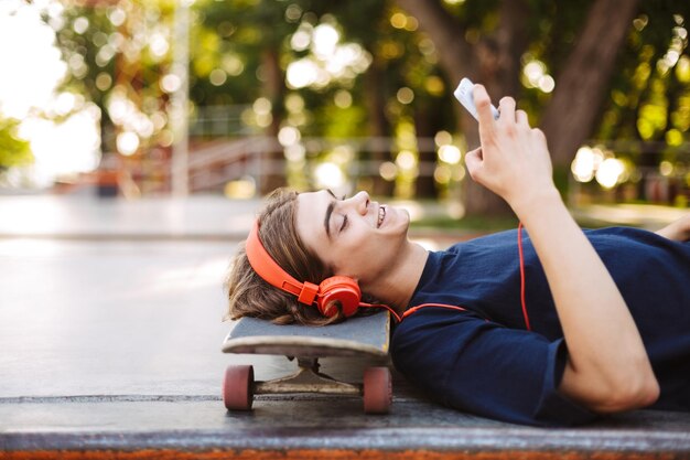 Portrait of young guy in orange headphones lying on skateboard while happily using cellphone spending time at skatepark