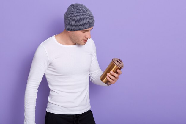 Portrait of young guy, standing and holding thermo mug, moving towards zero waste, posing on lilac, male wearing white casual shirt and gray cap. Copy space for advertisment.