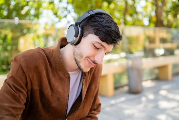 Портрет молодого красивого человека, слушающего музыку в наушниках, сидя на открытом воздухе. Городская концепция.
