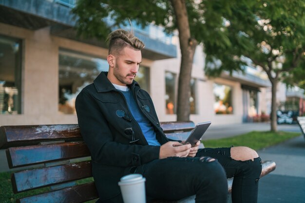 ベンチに座って屋外で彼のデジタルタブレットを使用して若いハンサムな男の肖像画。