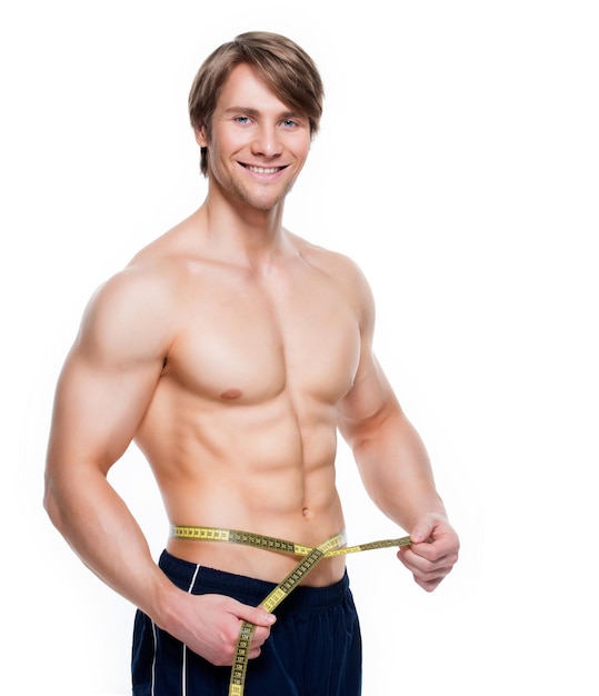 Free photo portrait of a young handsome man with muscular torso uses measuring tape on a white wall.