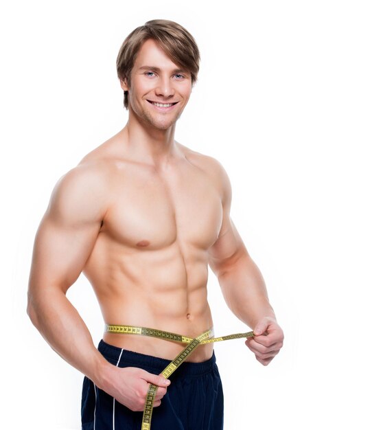 Portrait of a young handsome man with muscular torso uses measuring tape on a white wall.