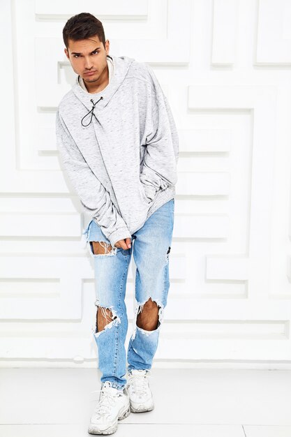 Portrait of young handsome model man dressed in gray casual hoodie clothes posing near textured white wall