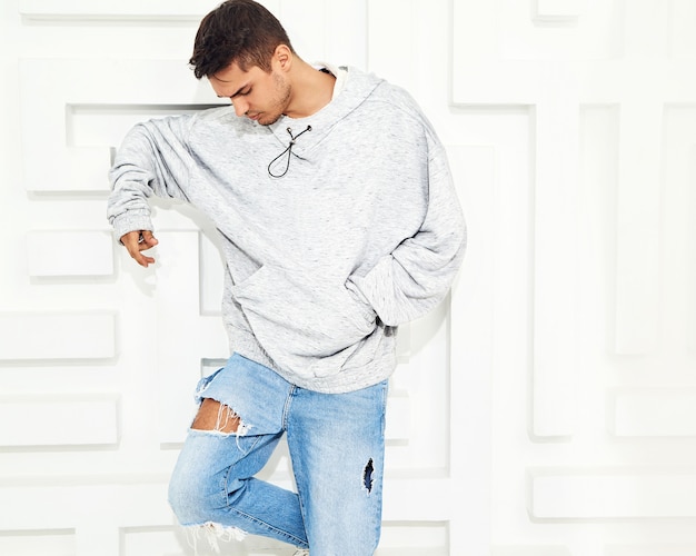 Portrait of young handsome model man dressed in gray casual hoodie clothes posing near textured white wall