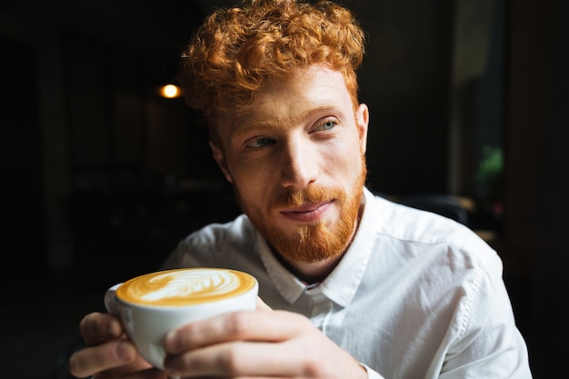 Portrait of young handsome redhead bearded man in white shirt holding coffee cup, looking aside