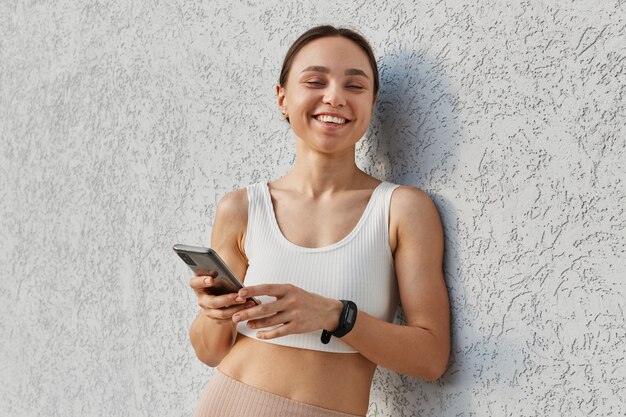 Portrait of young happy attractive brunette woman white top standing near gray wall and holding phone
