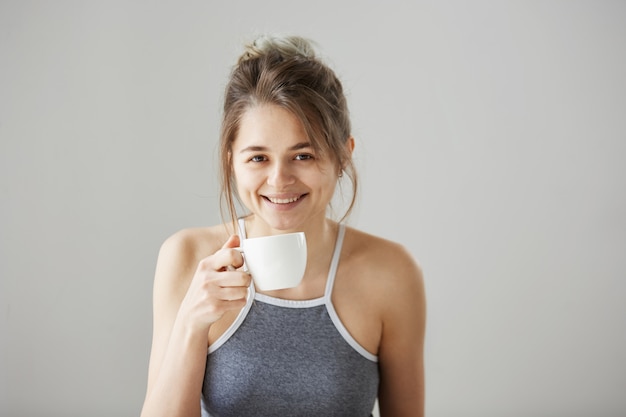 Портрет молодой счастливой красивой женщины усмехаясь держащ выпивая чашку кофе на утре над белой стеной.