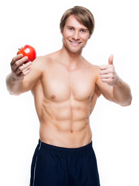 Portrait of young happy bodybuilder holding apple in his hand and show thumbs up sign - isolated on white wall.