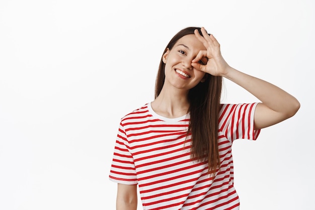 Portrait of young happy girl shows okay zero OK sign near eye and smiling tilt head carefree positive and joyful pose wearing summer tshirt white background