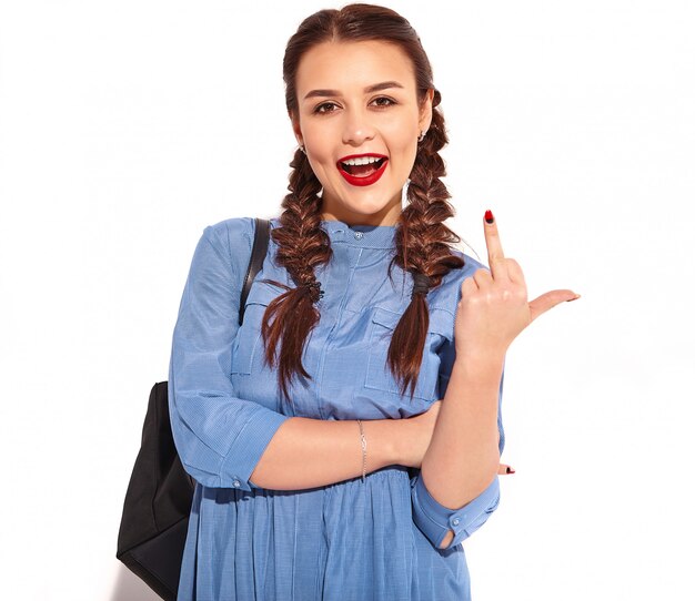 Portrait of young happy smiling woman model with bright makeup and red lips with two pigtails in hands in summer colorful blue dress and backpack isolated. Showing fuck off sign