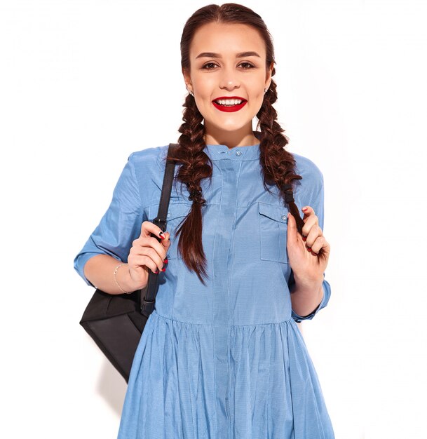 Portrait of young happy smiling woman model with bright makeup and red lips with two pigtails in hands in summer colorful blue dress and backpack isolated