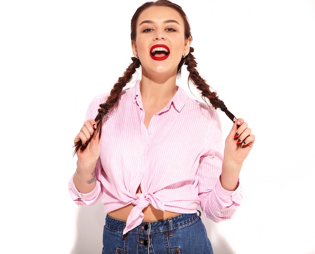Portrait of young happy smiling woman model with bright makeup and red lips with two pigtails in hands in summer colorful pink tied shirt isolated