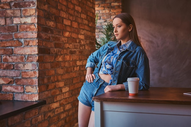 셔츠와 청바지 재킷, 반바지를 입은 젊은 힙스터 소녀의 초상화는 로프트 인테리어가 있는 방에서 테이크아웃 커피 한 잔과 함께 테이블에 기대어 있습니다.