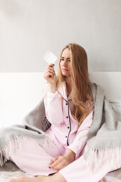 Portrait of young ill lady in sleepwear sitting with plaid on bed and thoughtfully looking on pills at home