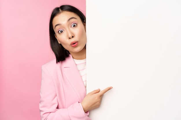 Free photo portrait of young japanese business woman corporate lady in suit pointing on wall with chart showing diagram or advertisement on empty copy space pink background