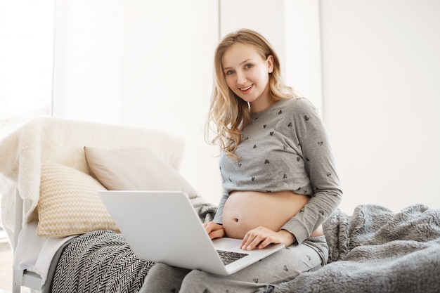 Портрет молодой радостной красивой беременной женщины с светлыми волосами в домашней одежде, сидя на кровати, улыбаясь, просматривая статьи о материнстве на портативном компьютере.