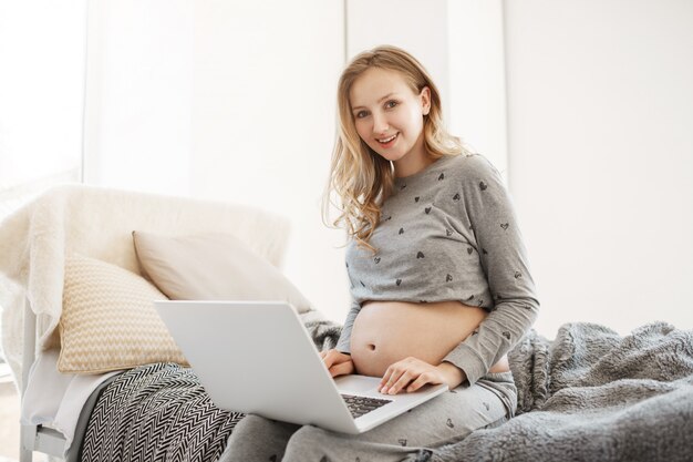 ラップトップコンピューターでの出産生活についての記事を見て、笑みを浮かべて、ベッドの上に座っているホームウェアで明るい髪の若いうれしそうな美しい妊娠中の女性の肖像画。