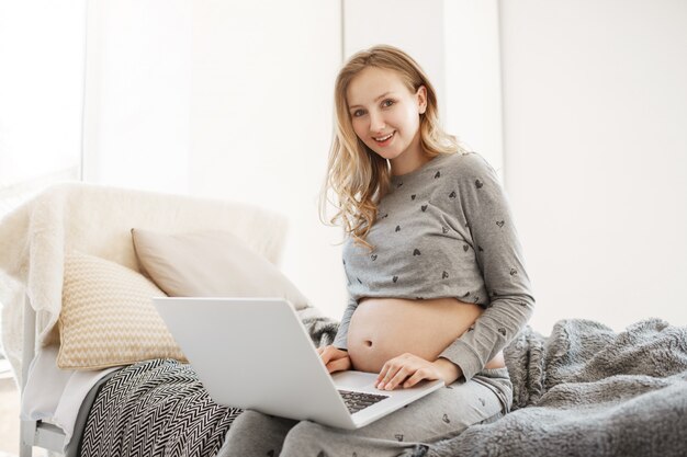 Portrait of young joyful beautiful pregnant woman with light hair in home wear sitting on bed, smiling, looking through articles about maternity life on laptop computer.