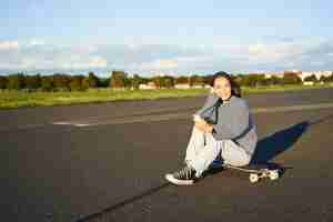 Free photo portrait of young korean girl sitting on her skateboard on road looking at smartphone chatting on mo