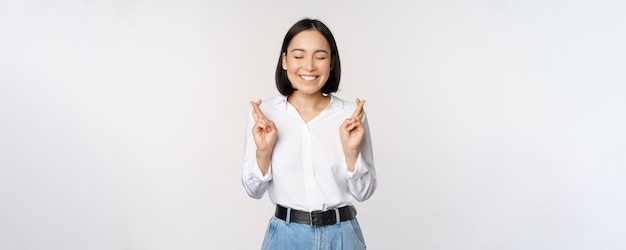 Portrait of young korean woman asian girl cross fingers and praying making wish anticipating waiting for results standing over white background