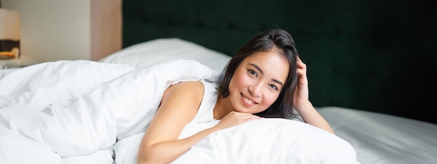 Free photo portrait of young korean woman smiling lying in bed on pillow posing in her bedroom looking relaxed