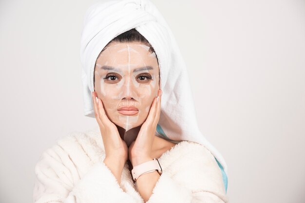 Portrait of young lady in bathrobe and towel on head while touching her face with facemask .