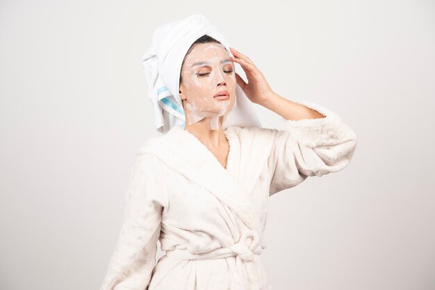 Portrait of young lady in bathrobe and towel on head while touching her face with facemask