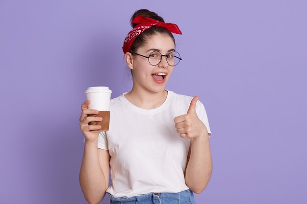 ライラックの空間に孤立したコーヒーのカップで立っているカジュアルな白いtシャツの若い女性の肖像画。美少女ながら楽しくジェスチャー親指とウィンクを示しています。