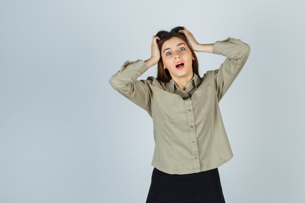 Portrait of young lady clasping head with hands in shirt, skirt and looking shocked front view