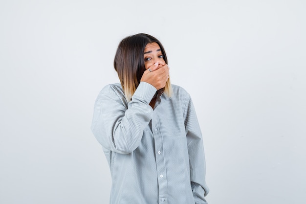 Portrait of young lady covering mouth with hand in oversized shirt and looking scared front view