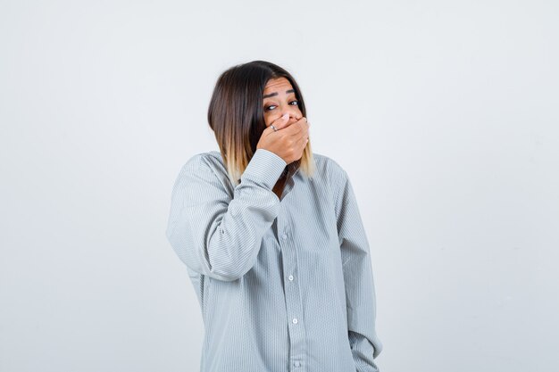 Portrait of young lady covering mouth with hand in oversized shirt and looking scared front view