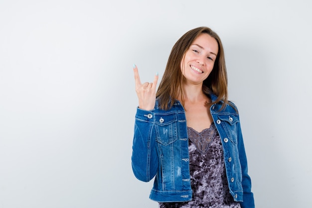 Portrait of young lady doing rock symbol in blouse, denim jacket and looking crazy front view