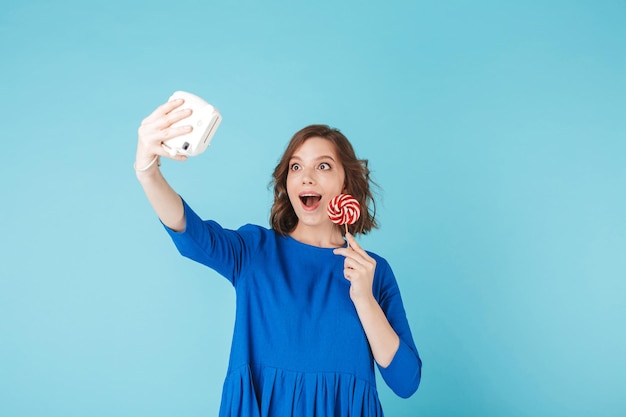 Portrait of young lady in dress standing with lollipop and taking cute photo on her little camera on over blue background