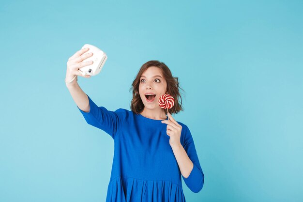 Portrait of young lady in dress standing with lollipop and taking cute photo on her little camera on over blue background