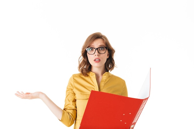 Portrait of young lady in eyeglasses standing with open red folder and amazedly looking in camera on white background isolated