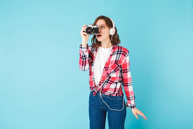ピンクの背景の上に写真を撮っている間、小さなレトロなカメラで驚いて見ているヘッドフォンの若い女性の肖像画
