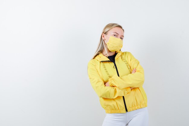 Portrait of young lady holding arms folded in jacket, pants, mask and looking proud front view