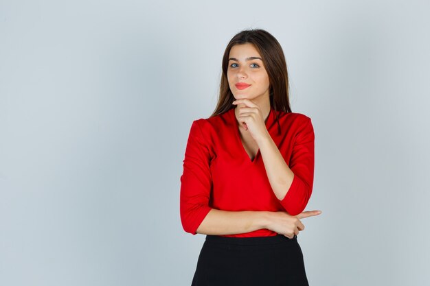 Portrait of young lady holding finger under chin in red blouse