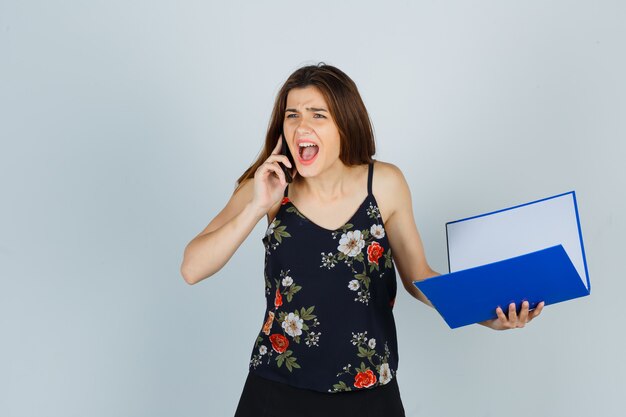 Portrait of young lady holding folder, talking on mobile phone while screaming in blouse and looking angry front view