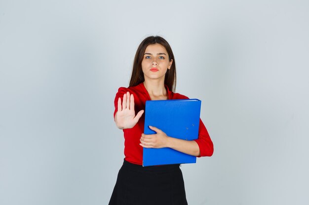 Portrait of young lady holding folder while showing stop gesture in red blouse