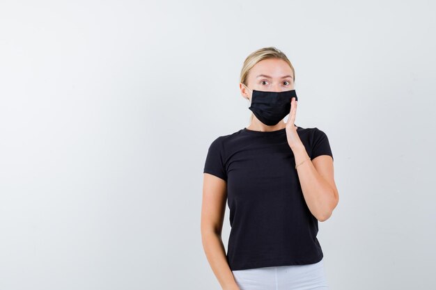 Portrait of young lady holding hand on mask in t-shirt, pants, medical mask