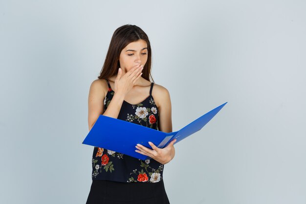 Portrait of young lady holding hand on mouth while looking over folder in blouse and looking surprised front view