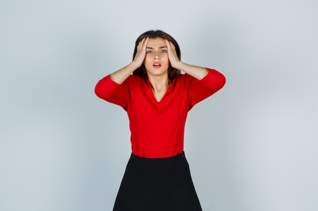 Free photo portrait of young lady holding hands on head in red blouse, skirt and looking forgetful