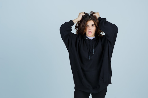 Portrait of young lady holding strand of hair in oversized hoodie, pants and looking fatigued front view