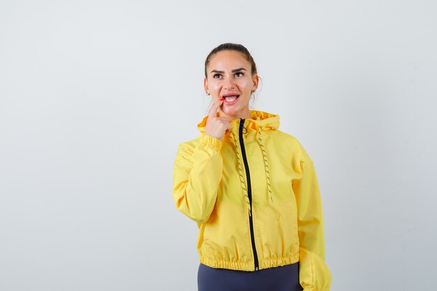 Portrait of young lady keeping finger on lip while looking up in yellow jacket and looking pensive front view