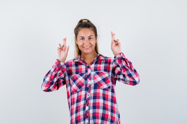 Portrait of young lady keeping fingers crossed in checked shirt and looking cheerful front view