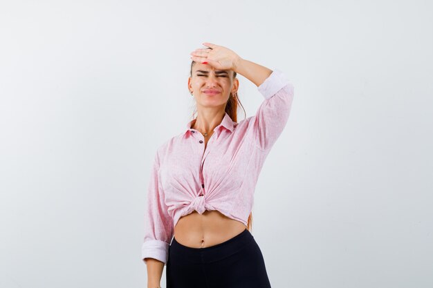 Portrait of young lady keeping hand on forehead in shirt, pants and looking forgetful front view