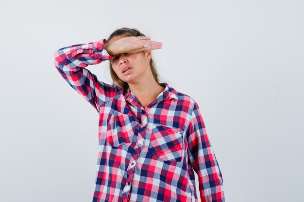 Portrait of young lady keeping hand over head in checked shirt and looking annoyed front view