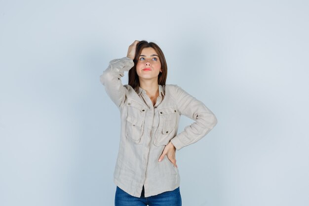 Portrait of young lady keeping hand on head while looking up in casual, jeans and looking pensive front view