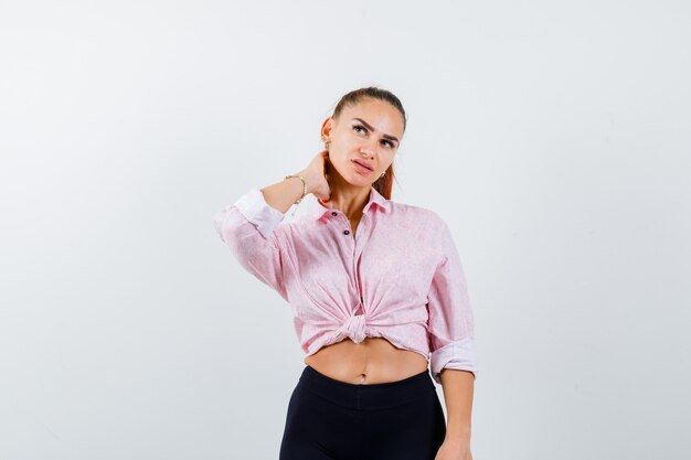 Portrait of young lady keeping hand on neck in shirt, pants and looking pensive front view